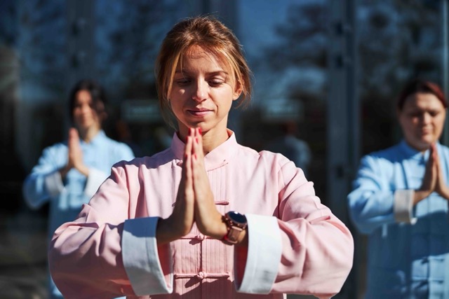 Quest for Balance Wellness offers Tai chi. Pictured, a young woman is smiling while meditating outdoors on a sunny day with her qigong group.